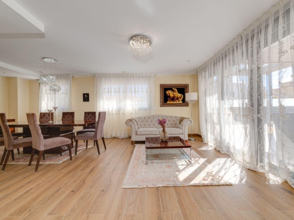 Open living and dining area with a wooden dining table and eight mauve chairs, lace curtains, and a tufted beige sofa in a bright space.