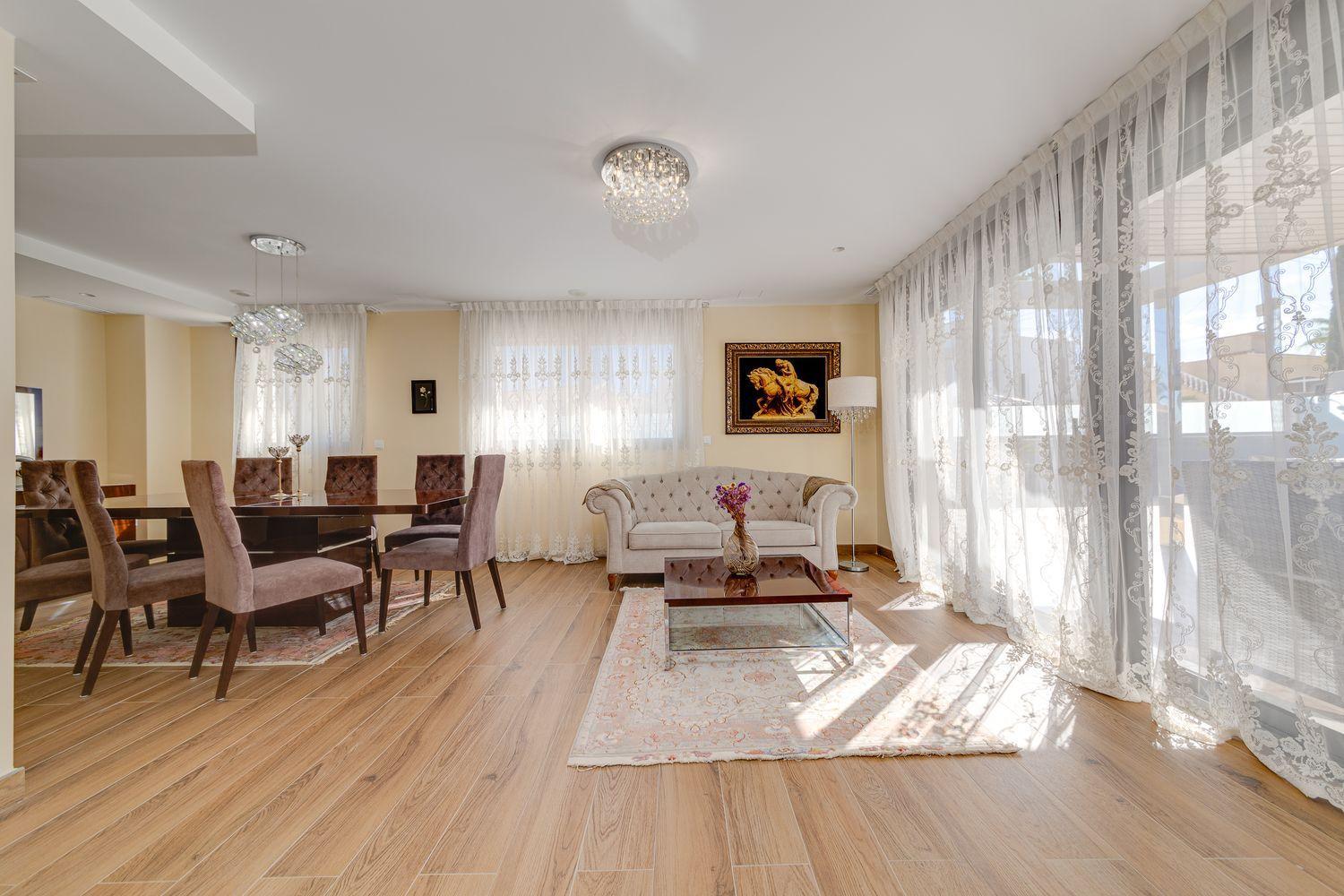 Open living and dining area with a wooden dining table and eight mauve chairs, lace curtains, and a tufted beige sofa in a bright space.