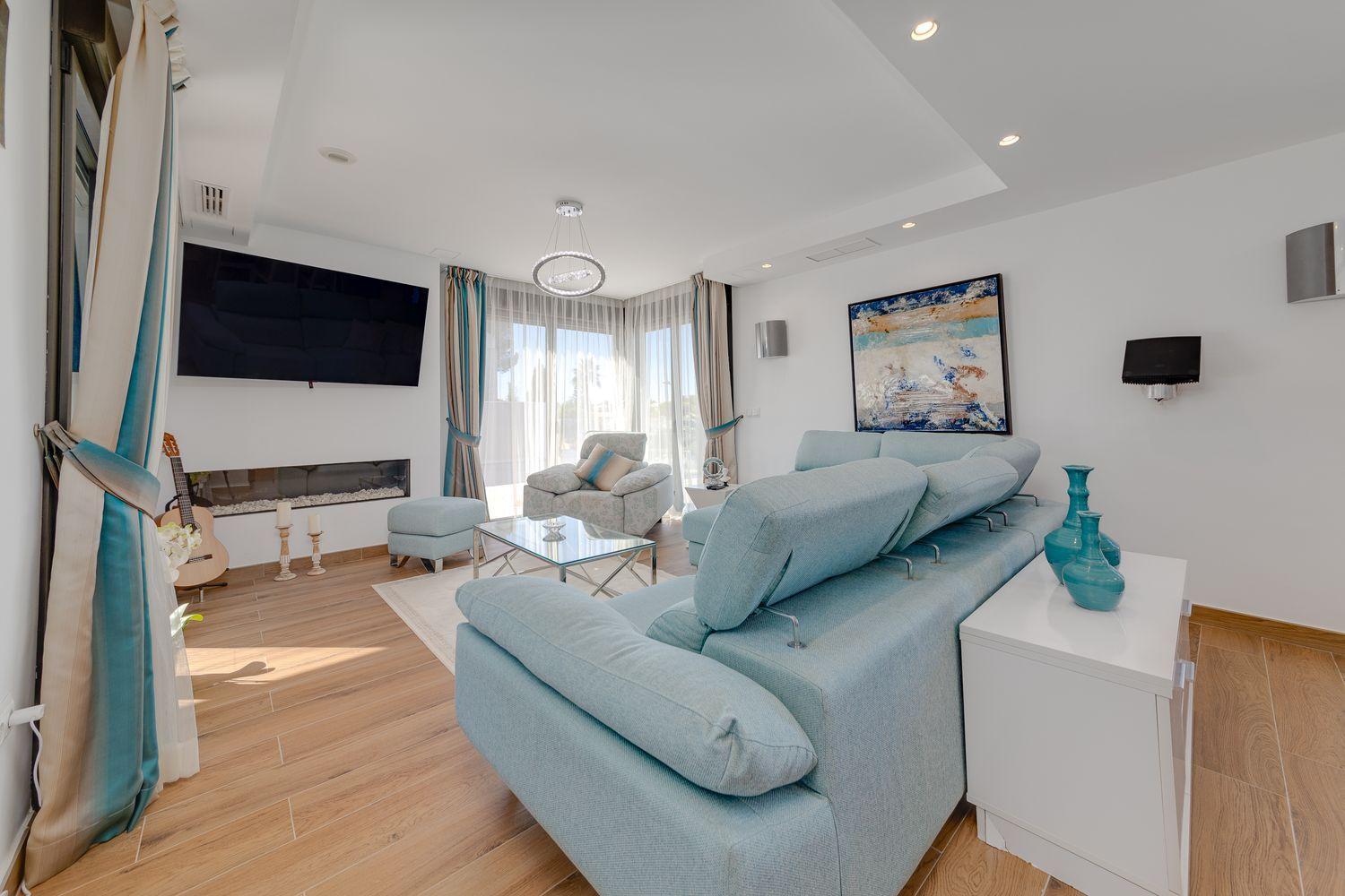 Bright living room with a light-blue sectional sofa, glass coffee table, and a wall-mounted TV above a linear fireplace.