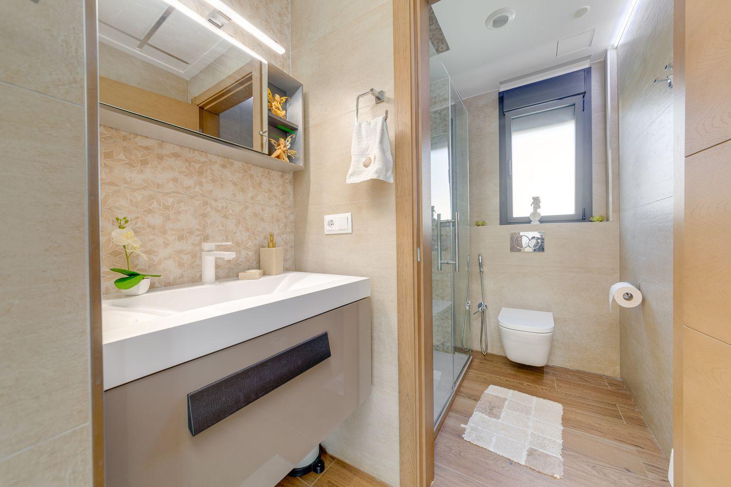 Modern bathroom with beige tiles, white sink vanity, wall mirror, glass shower, and wall-mounted toilet.