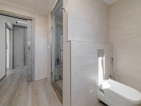 Contemporary bathroom with a wall-mounted toilet, handheld bidet spray, and a glass shower enclosure; beige tile walls and wood-look flooring.