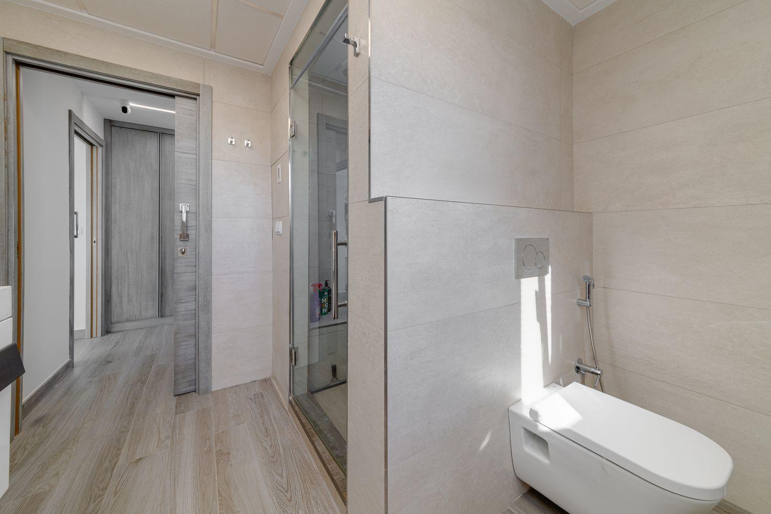 Contemporary bathroom with a wall-mounted toilet, handheld bidet spray, and a glass shower enclosure; beige tile walls and wood-look flooring.