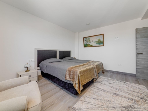 Bedroom with a double bed, gray bedding, and two matching nightstands, a small lamp, framed landscape painting, and a beige armchair in the corner.