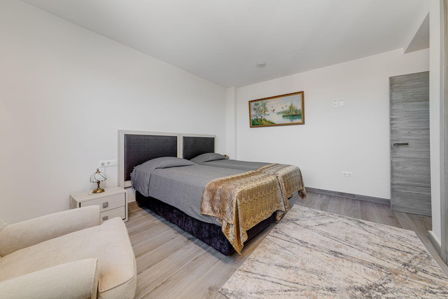 Bedroom with a double bed, gray bedding, and two matching nightstands, a small lamp, framed landscape painting, and a beige armchair in the corner.