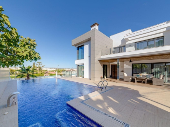 Modern white, multilevel villa with a large rectangular pool and glass doors opening to a shaded outdoor seating area on a sunny day.