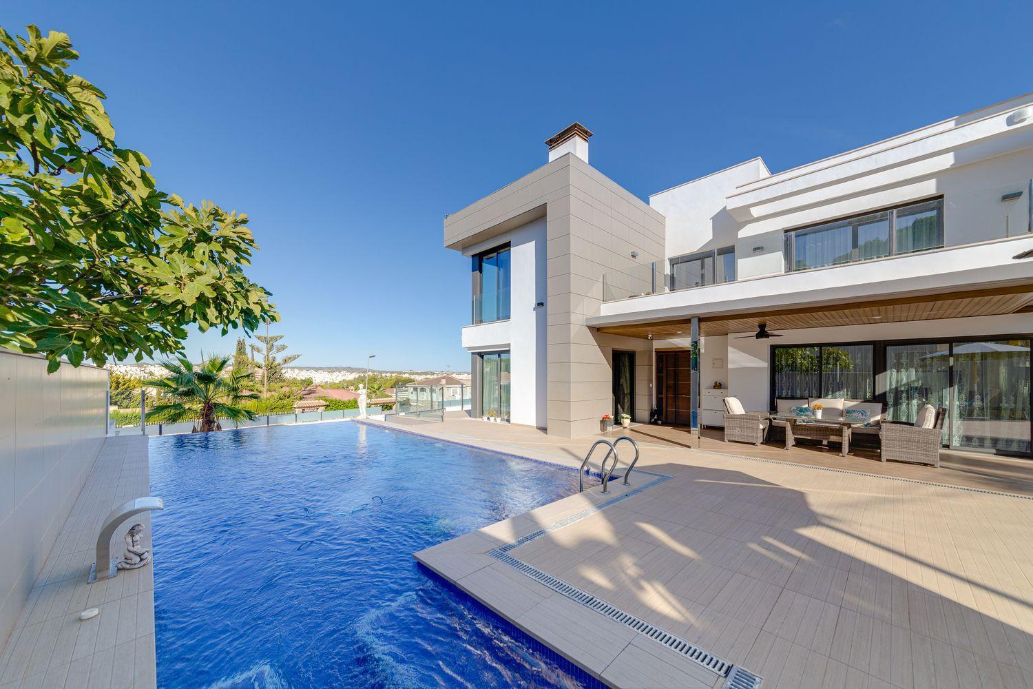 Modern white, multilevel villa with a large rectangular pool and glass doors opening to a shaded outdoor seating area on a sunny day.
