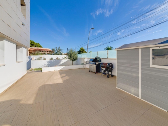 Sunlit courtyard with beige tiled patio, white building on left and gray shed on right; a barbecue grill nearby.