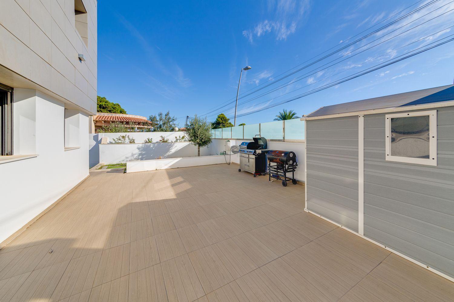 Sunlit courtyard with beige tiled patio, white building on left and gray shed on right; a barbecue grill nearby.