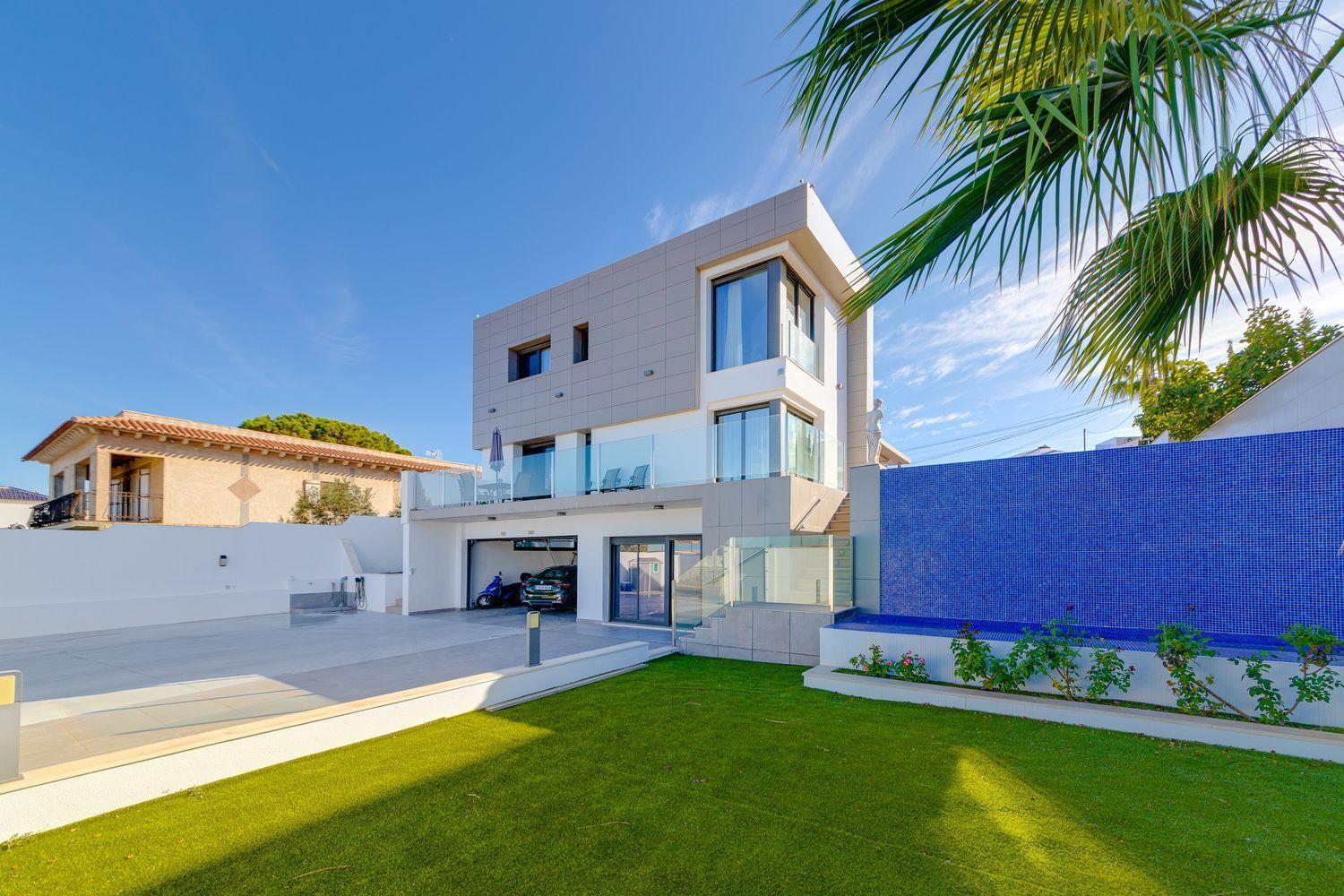 Modern white multi‑story house with glass balconies, a blue tiled wall, and a green lawn under a clear sky.