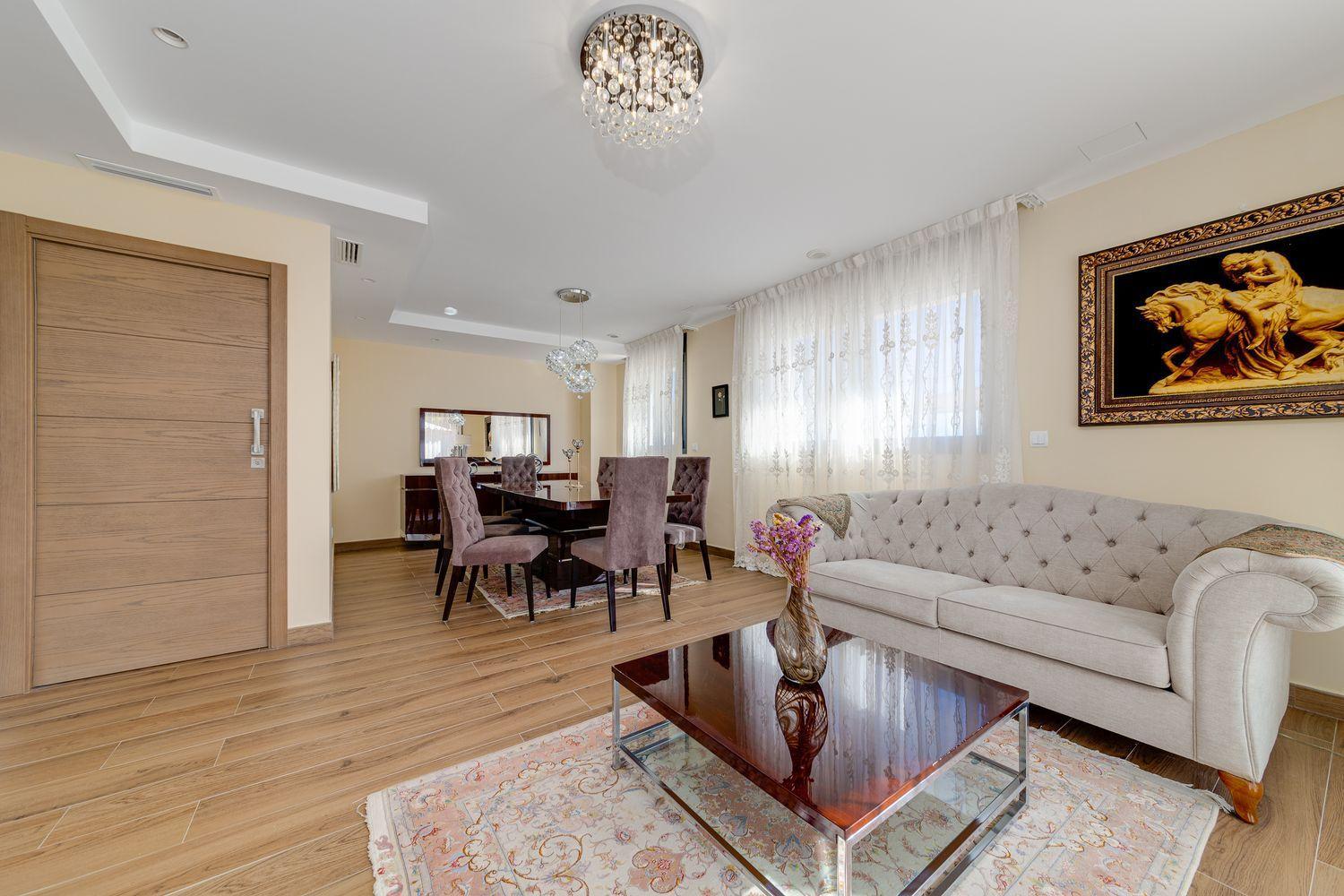 Open living and dining room with a beige tufted sofa, dark wood dining table, and a glass coffee table on a warm wood floor