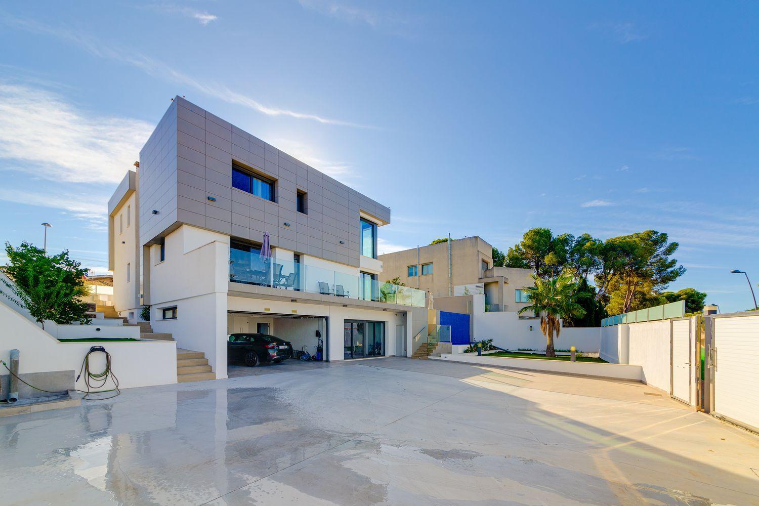 Modern white three-story house with glass balcony and driveway, blue sky in the background
