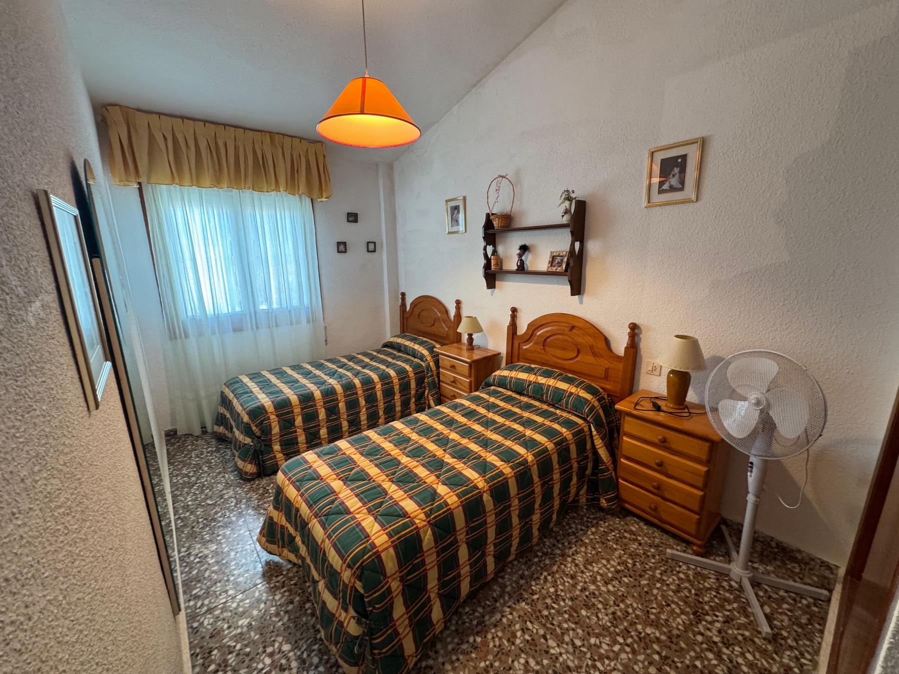 Twin bedroom with two single beds in plaid green, yellow, and red bedding, wooden headboards, and an orange pendant light overhead.