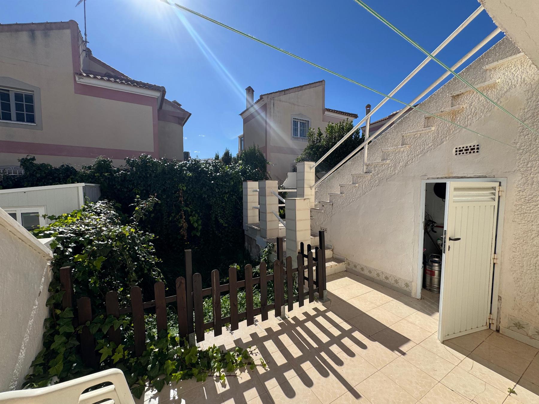 Sunny outdoor courtyard with beige houses, a tiled patio, and a wooden fence; sun glare creates long shadows from overhead clotheslines and a stairway on the right.