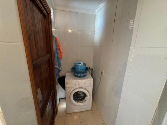 Narrow laundry room with a front-loading washing machine, blue basin on top, and towels hanging on the wooden door to the left.
