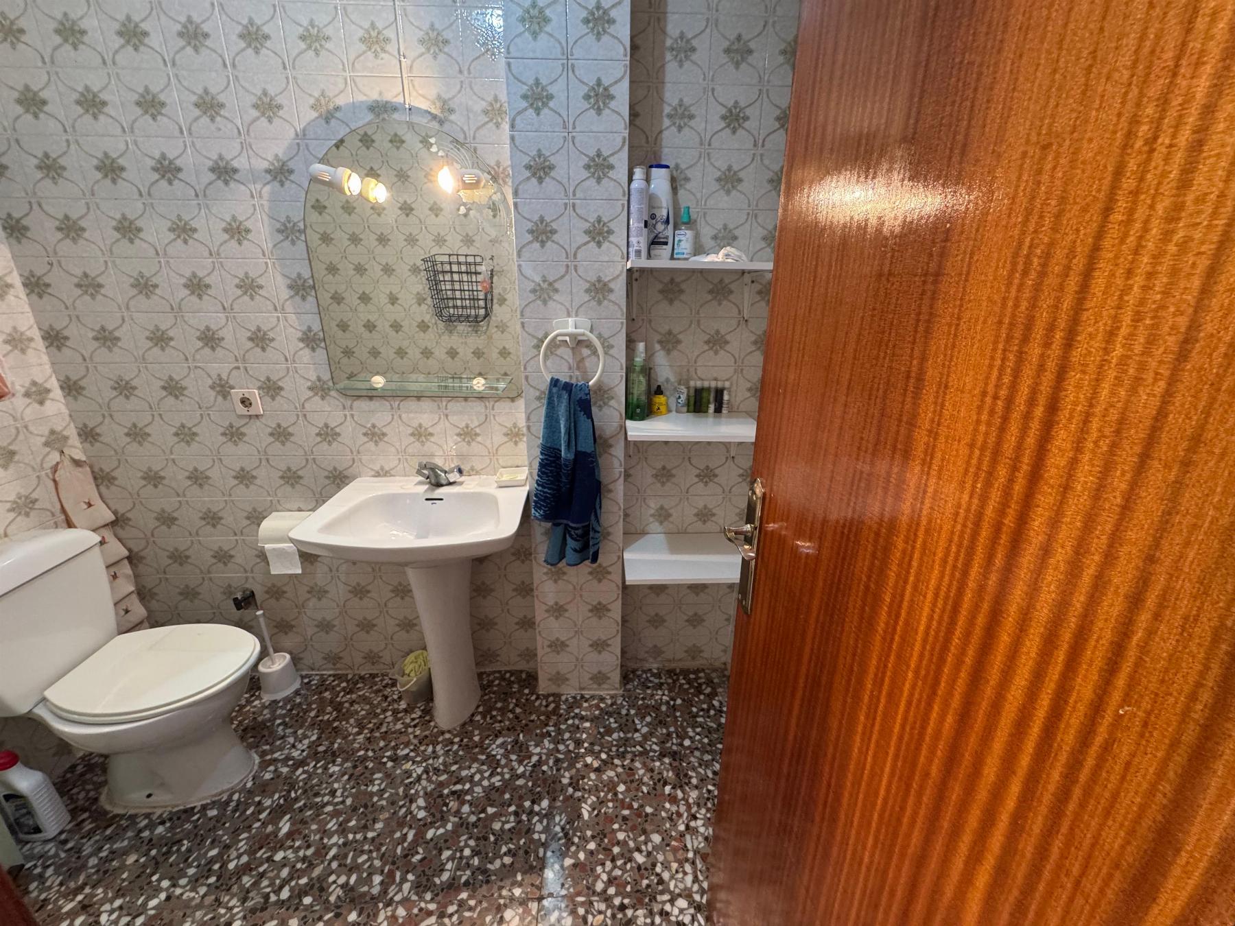 Small bathroom with patterned tile walls, pedestal sink, oval mirror, toilet, blue towel, and open shelf with toiletries.