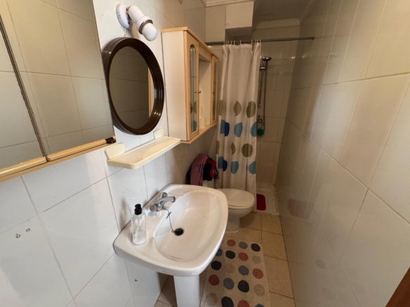 Small bathroom with white sink, round mirror, wall cabinet, toilet, and a shower with a polka-dot curtain.