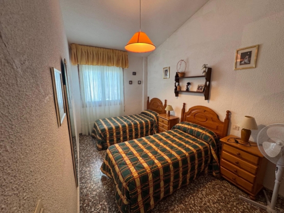 Twin bedroom with two single beds, green and yellow plaid bedspreads, wooden headboards, and matching nightstands under a warm ceiling light.