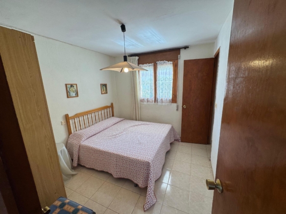 Twin-bed bedroom with patterned bedspreads, wooden headboard, and a hanging ceiling light; window with lace curtains on the far wall