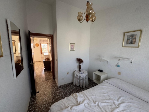 Bedroom corner with a white quilted bed, a small round table with a vase of dried flowers, wall art, and a hanging chandelier; doorway to a tile-floored hallway.
