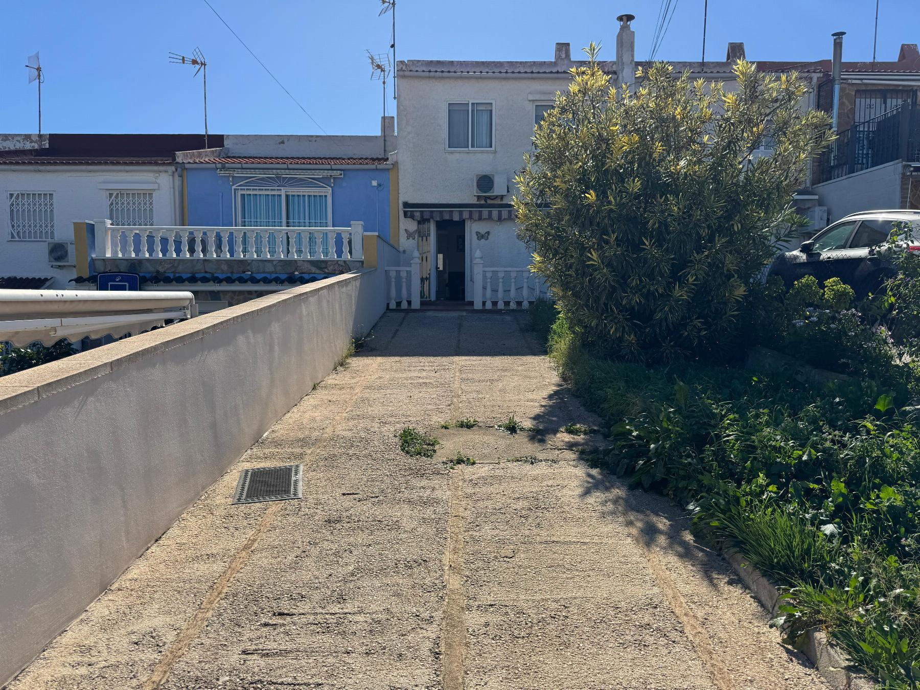 Driveway leading to a white two‑story house with a blue front facade and white railing, bordered by a large shrub on the right under a clear blue sky.