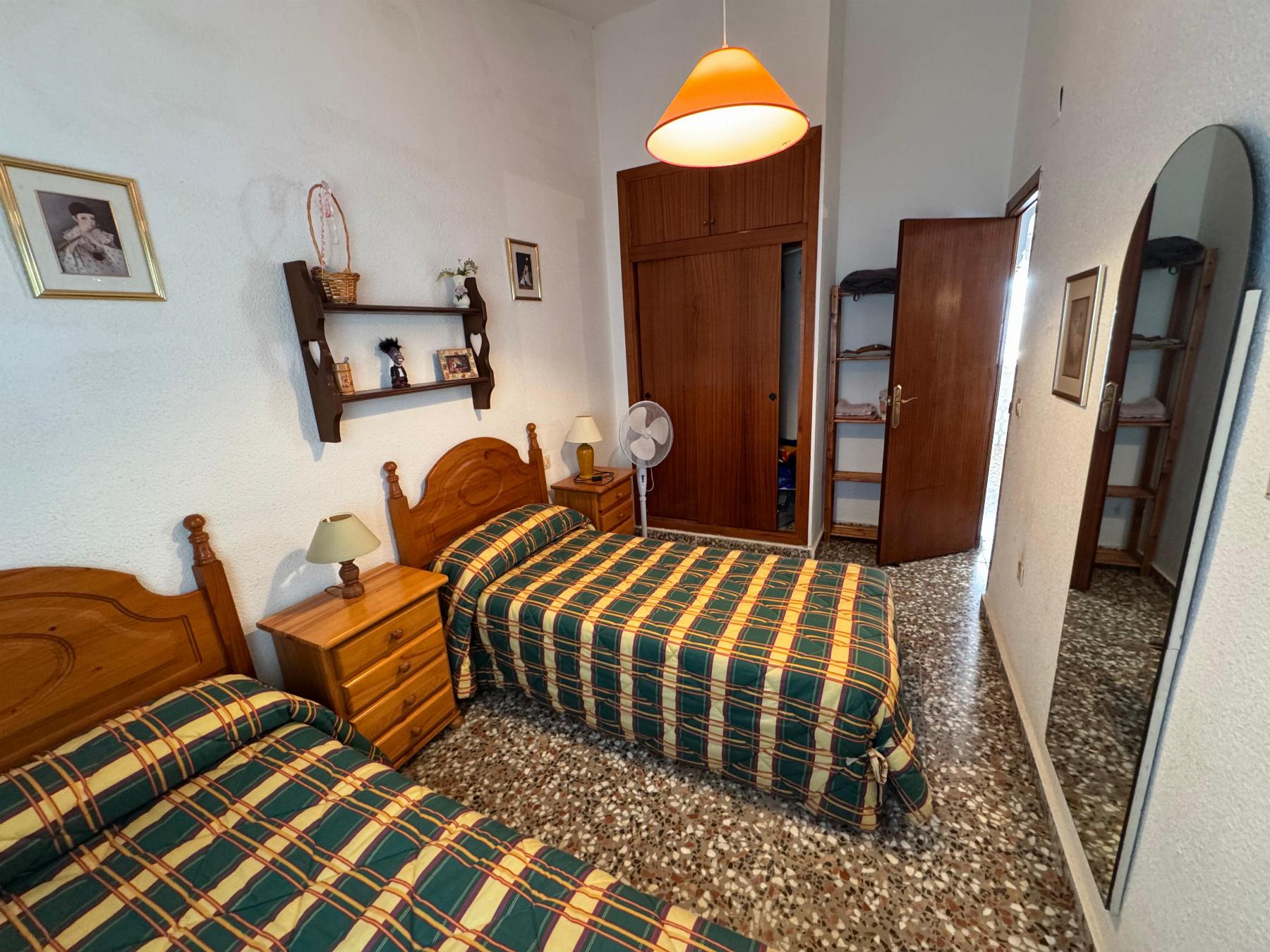 Twin bedroom with two wooden single beds and plaid green-yellow bedspreads, matching nightstands, and a wall shelf with decor.