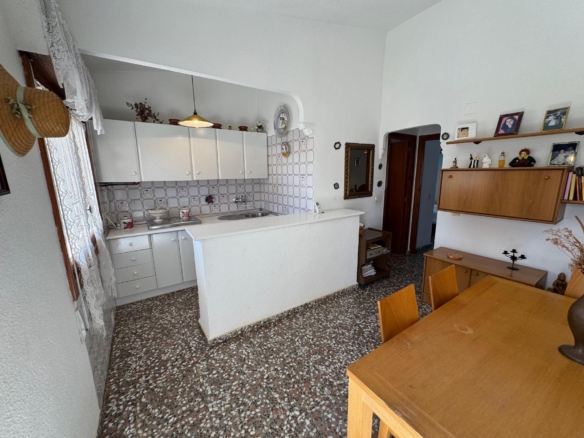 Bright kitchen with white cabinets, tiled backsplash, double sink, and a pass-through opening to a dining area with a wooden table and shelves of photos and decor.