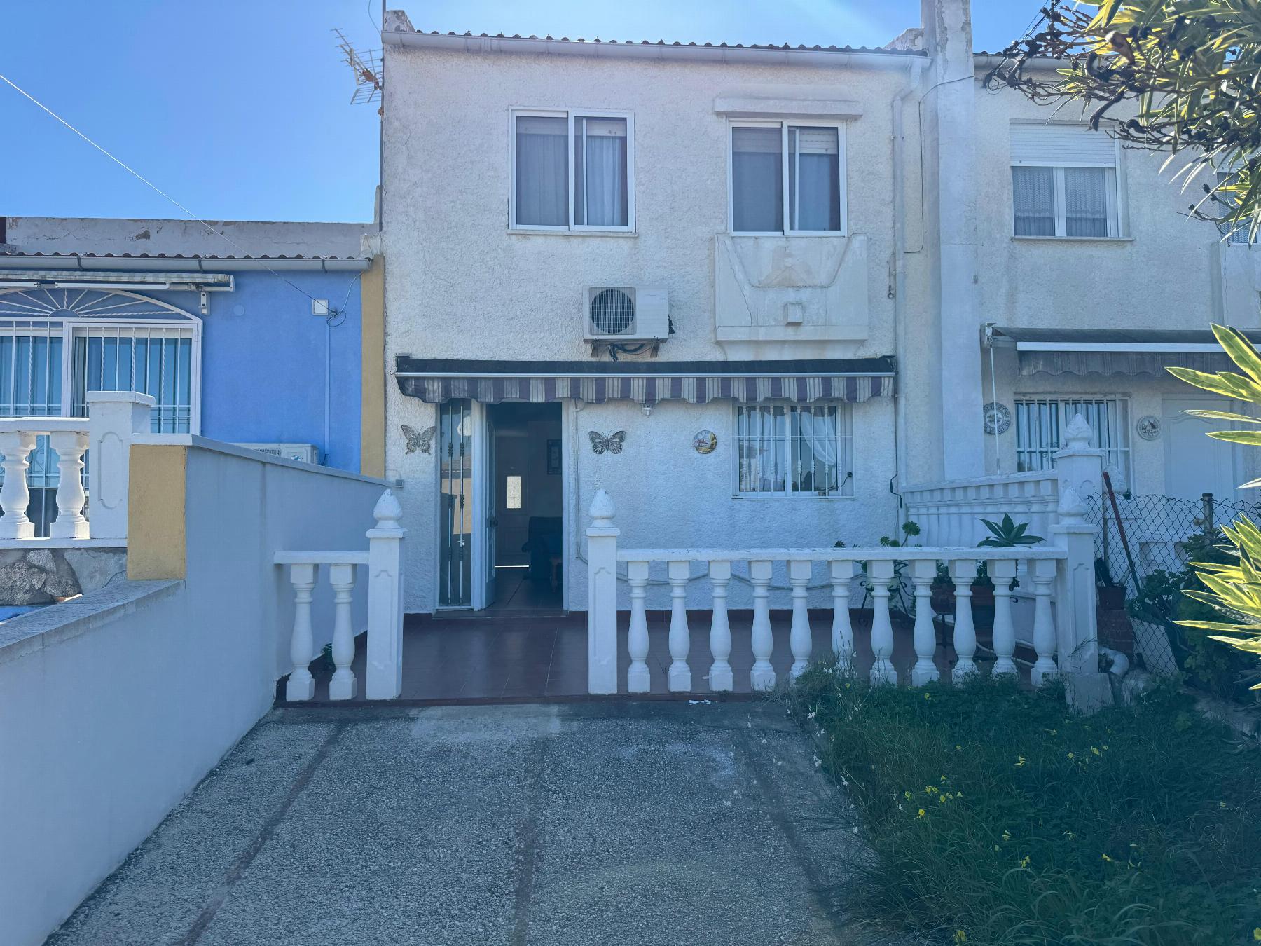 White two-story house with a blue side wall, barred windows, and butterfly decorations near the entrance; tiled walkway and white rail fence in front.