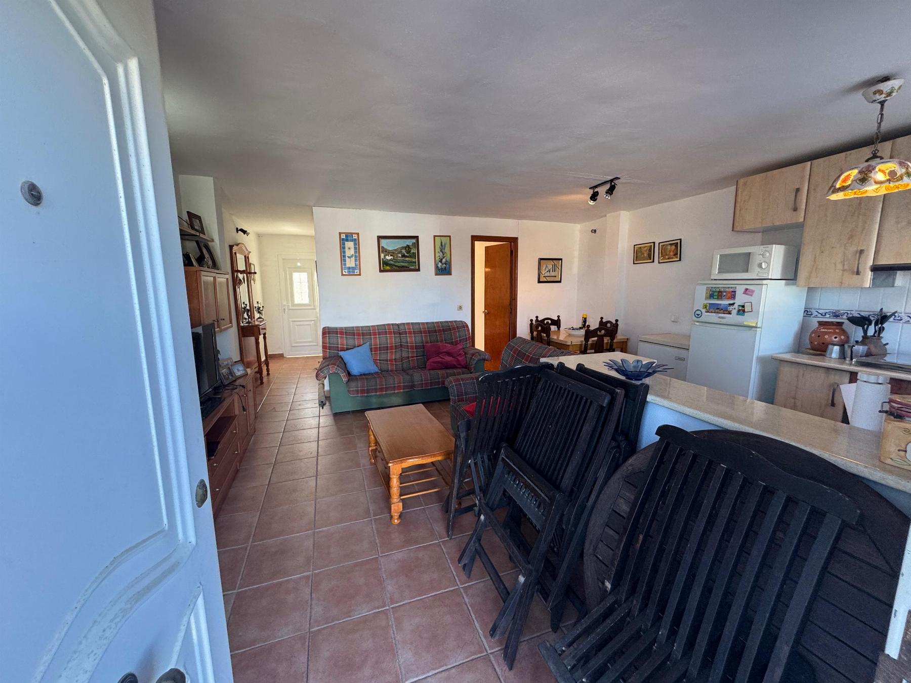 Cozy living room with plaid sofa, blue and red cushions, wooden coffee table, and wall art; kitchen area visible to the right and stacked chairs in front.