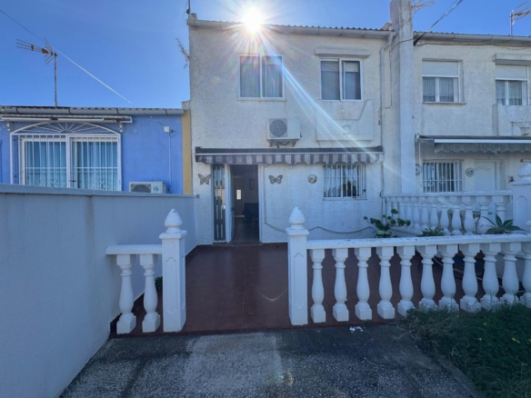 White two-story stucco house with a brown-tiled front walkway, white decorative railing, and a sun flare overhead; a blue neighboring house is on the left.