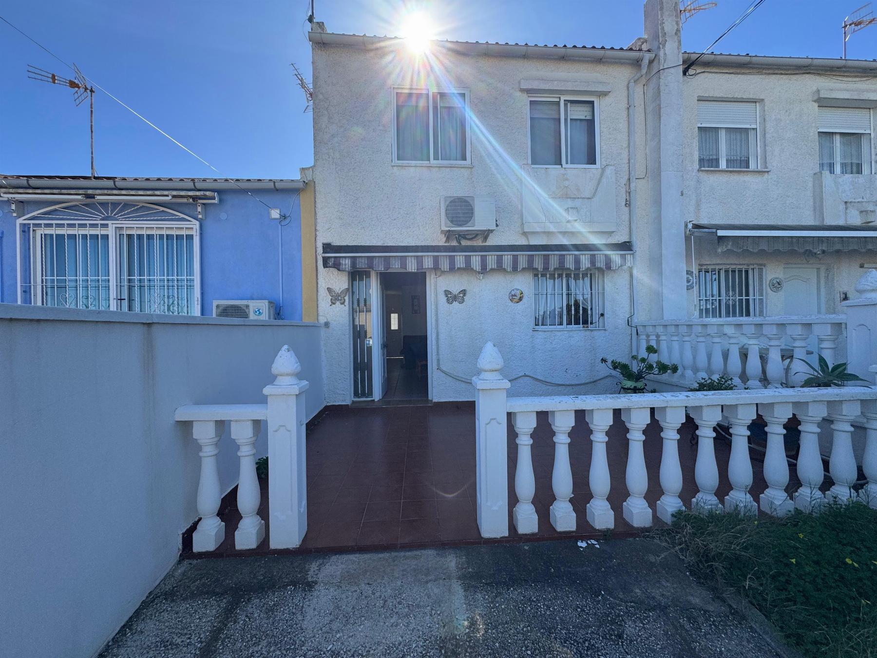 White two-story stucco house with a brown-tiled front walkway, white decorative railing, and a sun flare overhead; a blue neighboring house is on the left.