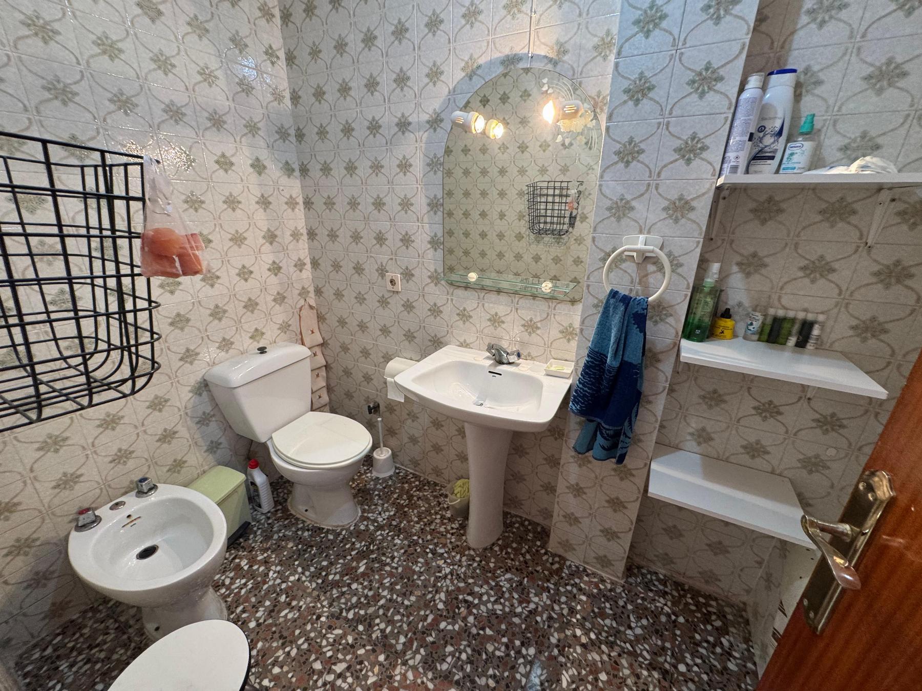 Small bathroom with patterned tile walls, toilet, bidet, and pedestal sink beneath a curved mirror and shelf storage.