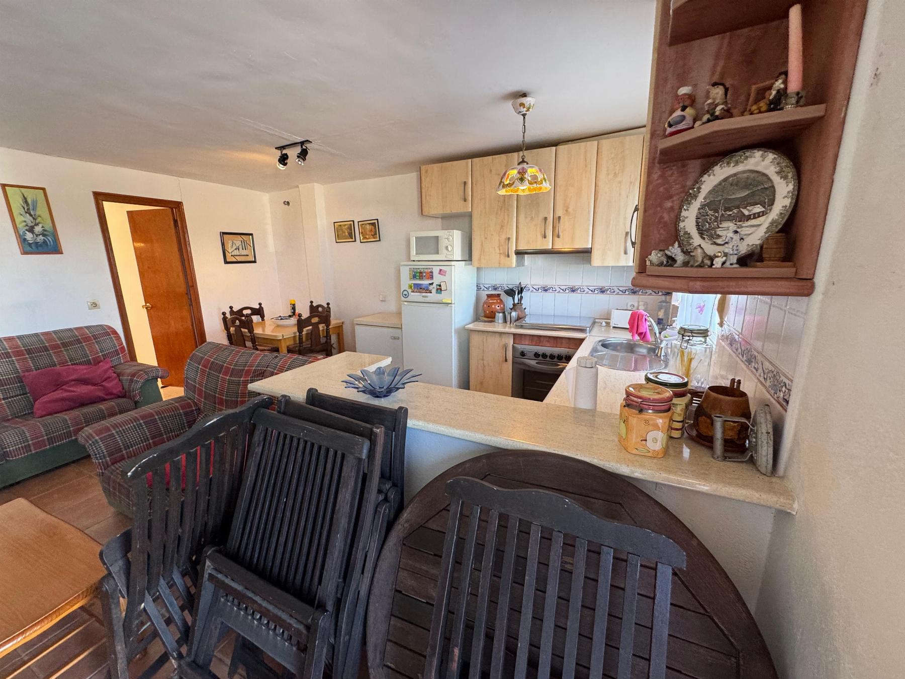 Open-plan kitchen and living area with plaid sofas, a small dining table, and stacked black chairs in the foreground.