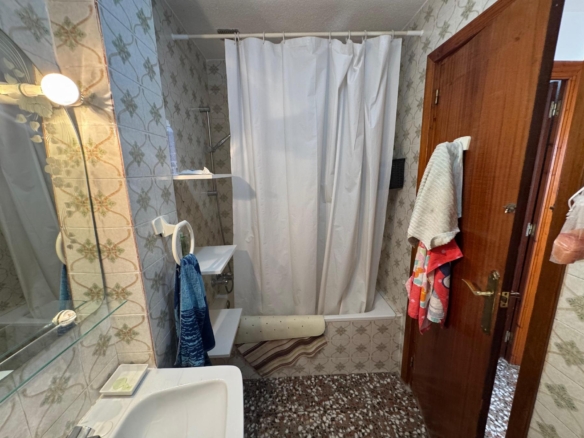 Small vintage bathroom with patterned tiles, white shower curtain, and towels by a wooden door.