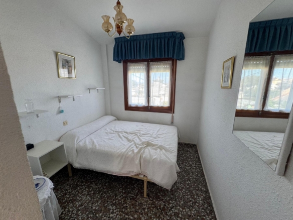 Cozy bedroom with white textured walls, a double bed in white linens, blue valance above a wooden-framed window, a hanging chandelier, and a mirrored wall nearby.