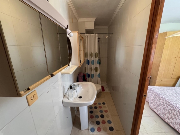 Small tiled bathroom with a pedestal sink, wall mirror cabinet, and a polka-dot shower curtain; doorway to a bedroom beyond the shower stall.
