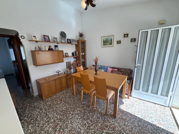 Dining area with a wooden table and chairs, wall shelves with framed photos, and a glass-front cabinet; a cushioned bench along the wall and sliding glass doors on the right.