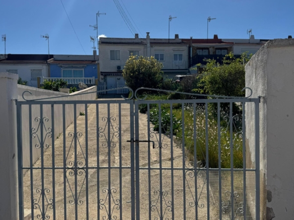 Blue sky above a gated courtyard; metal gate in foreground leads to a row of white townhouses and greenery beyond.