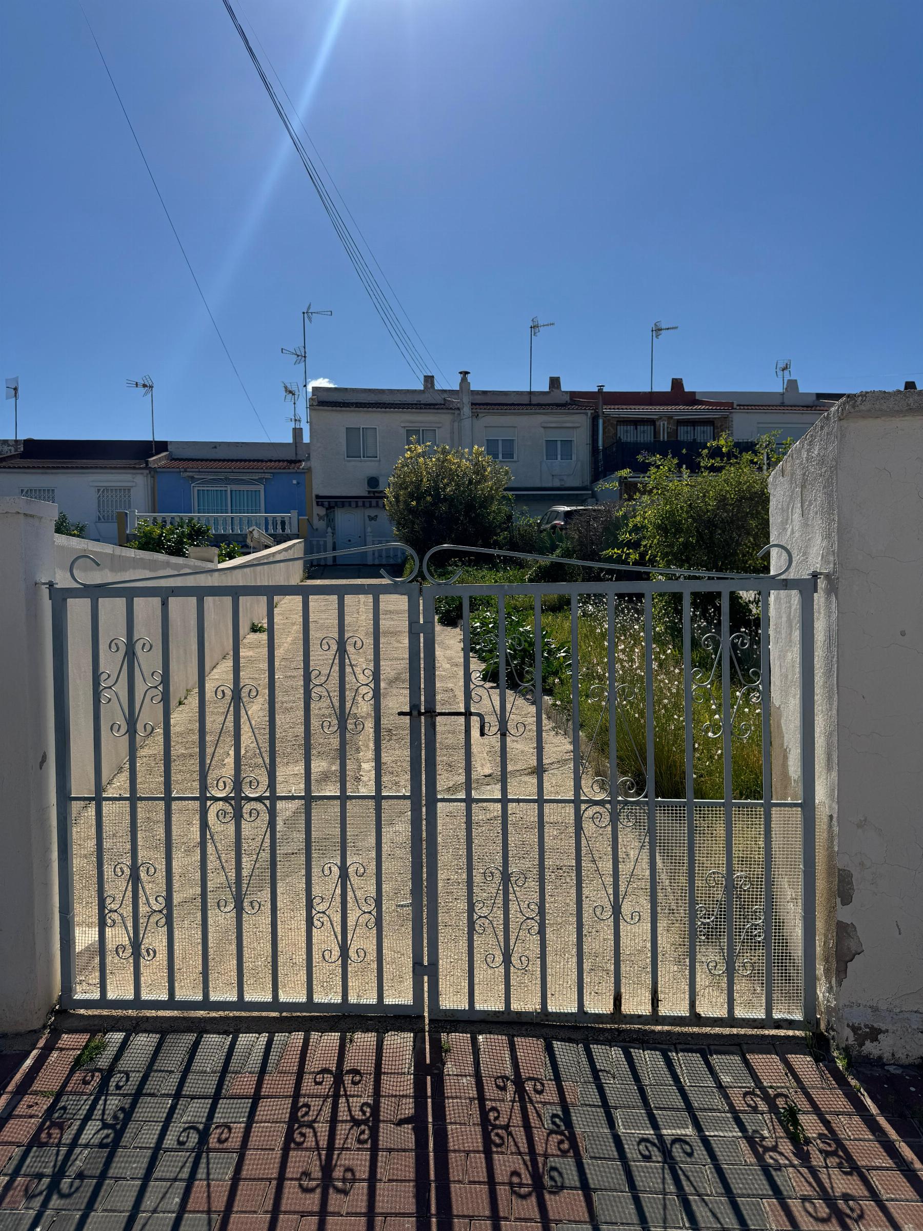 Blue sky above a gated courtyard; metal gate in foreground leads to a row of white townhouses and greenery beyond.