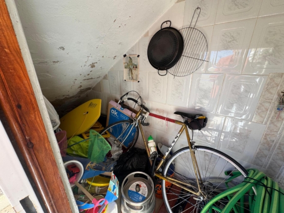 Cluttered storage area with a bicycle among pans, hoses, and boxes against a tiled wall under a staircase.