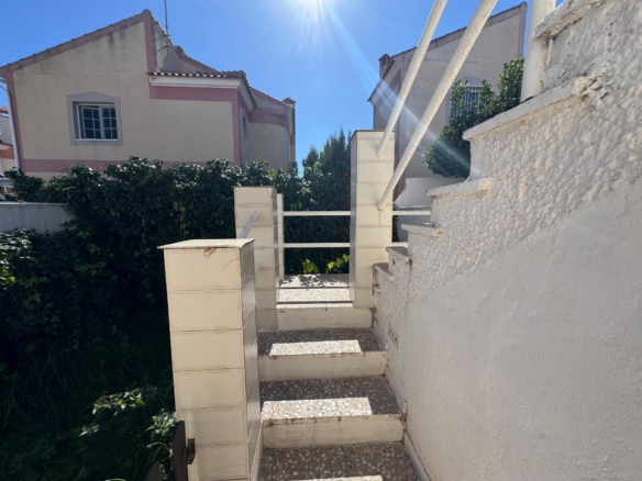 Sunlit outdoor stairway with white walls, beige mailbox towers, and green hedge along a Mediterranean-style home garden.