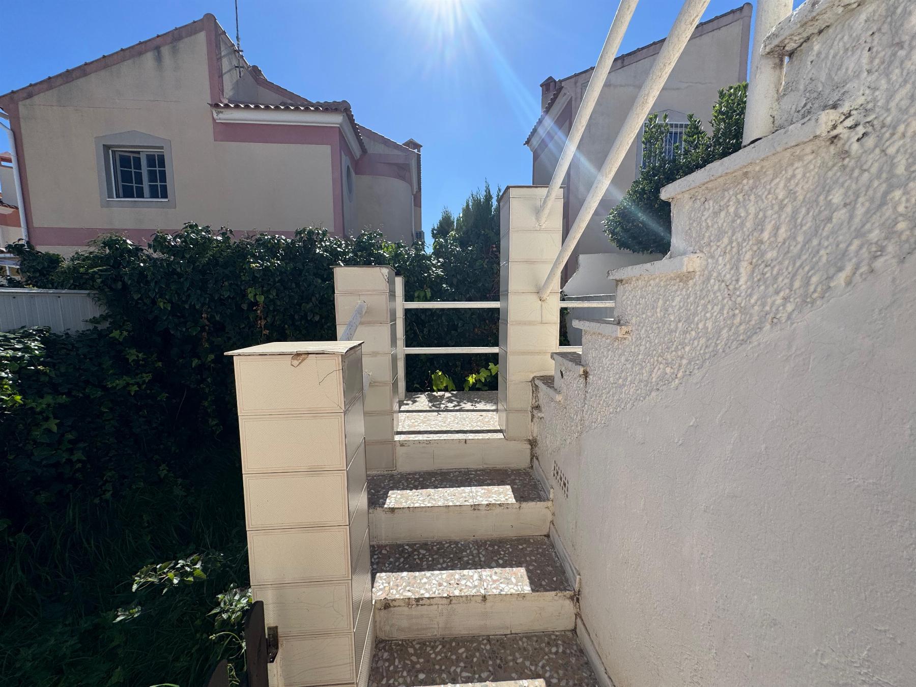 Sunlit outdoor stairway with white walls, beige mailbox towers, and green hedge along a Mediterranean-style home garden.