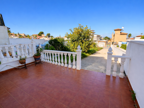 Terracotta-tiled patio with white balustrade and potted plants, overlooking a sunny Mediterranean neighborhood.