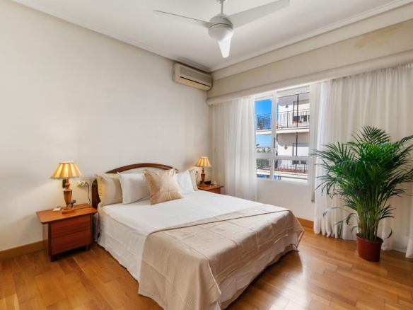 Bright hotel-style bedroom with a white bed flanked by wooden nightstands and lamps, large window with sheer curtains, ceiling fan, wall-mounted AC, and a potted palm by the window.