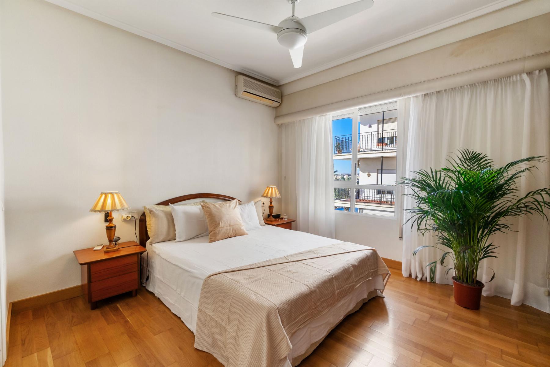 Bright hotel-style bedroom with a white bed flanked by wooden nightstands and lamps, large window with sheer curtains, ceiling fan, wall-mounted AC, and a potted palm by the window.