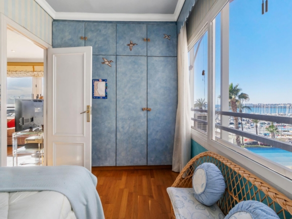 Bright seaside bedroom with blue wardrobe and wicker chair, large window showing a marina and palm trees outside.