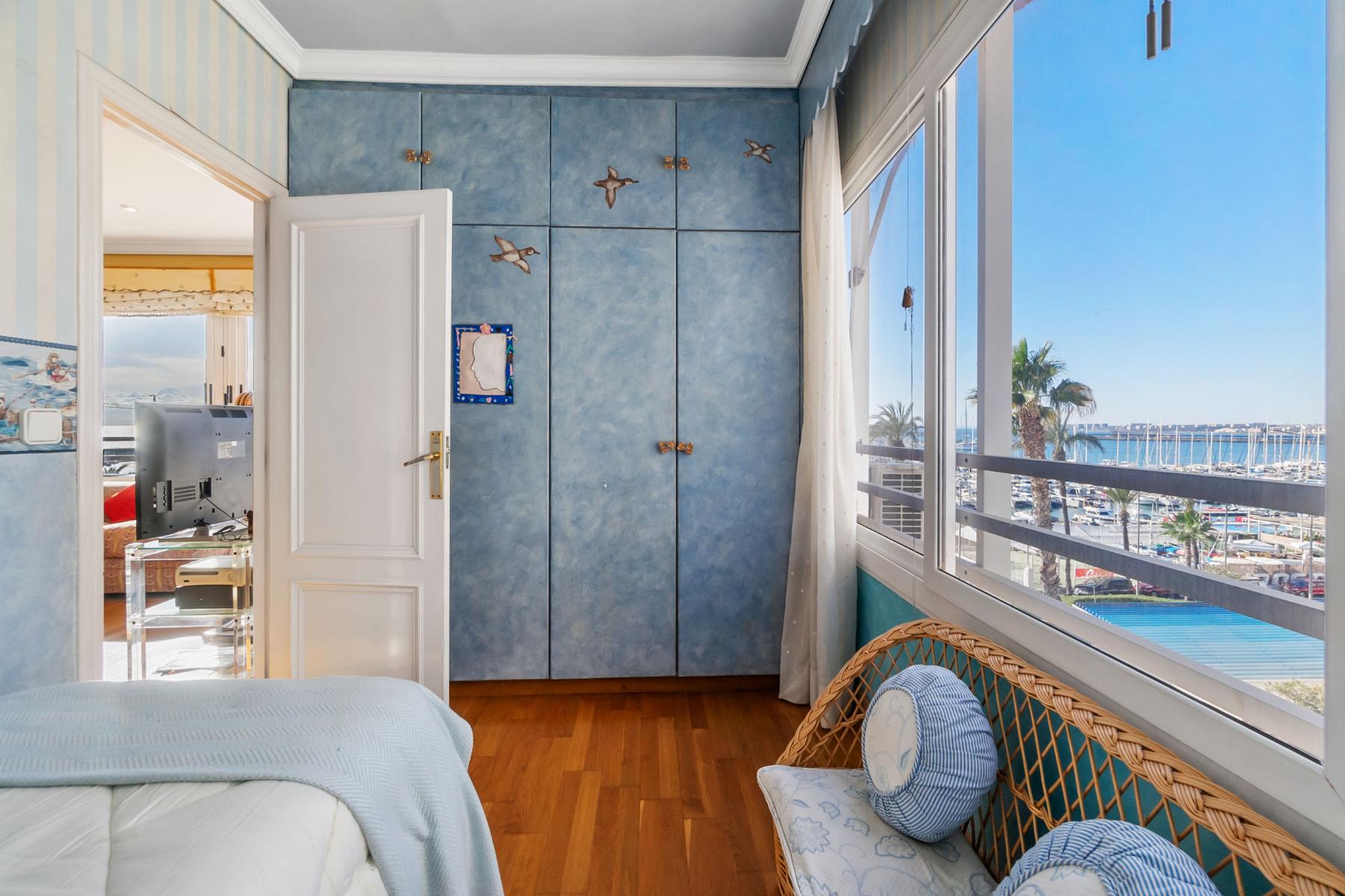 Bright seaside bedroom with blue wardrobe and wicker chair, large window showing a marina and palm trees outside.