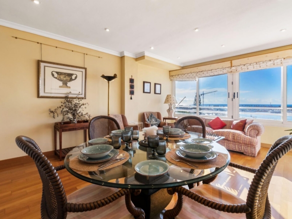 Bright coastal dining room with a round glass table set for six, wicker chairs, and large windows showing the sea view.