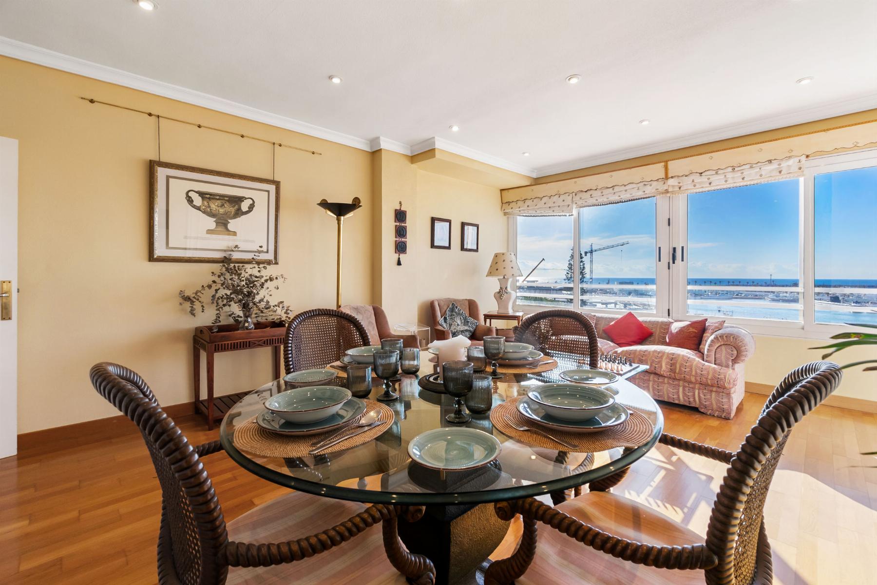 Bright coastal dining room with a round glass table set for six, wicker chairs, and large windows showing the sea view.