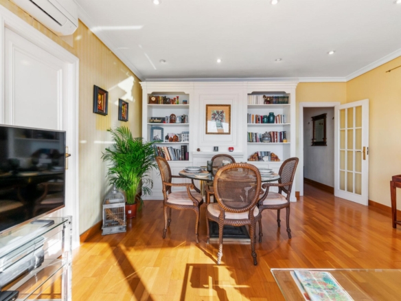 Bright living-dining room with white built-in cabinets, round glass table, wicker chairs, and warm yellow walls. Linear hardwood floor.