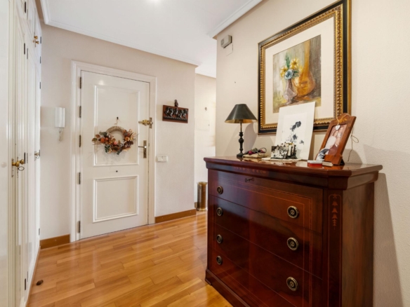 Hallway with white doors on left, a wreath on the front door, and a polished wooden dresser against the right wall topped with a lamp and framed art.